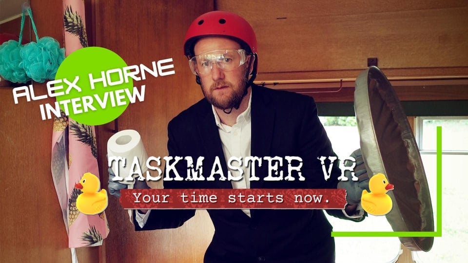 Alex Horne in Taskmaster holding a toilet roll and a garbage can lid while wearing a helmet.