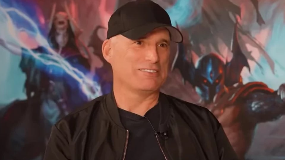 A photo of Christopher Perkins sitting in front of a painting of an adventuring party.